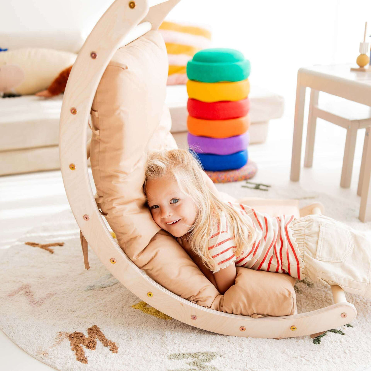 Kind liegt spielend auf dem Kletterbogen aus Holz in einem hellen Raum, umgeben von bunten Kissen.